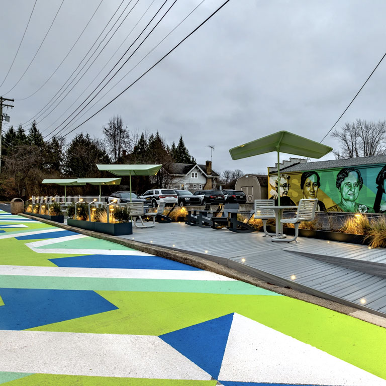 A large geometric mural painted in bright green, blue and white on asphalt, next to a mural of portraits of pioneering women from Salem, Indiana in similar colors.