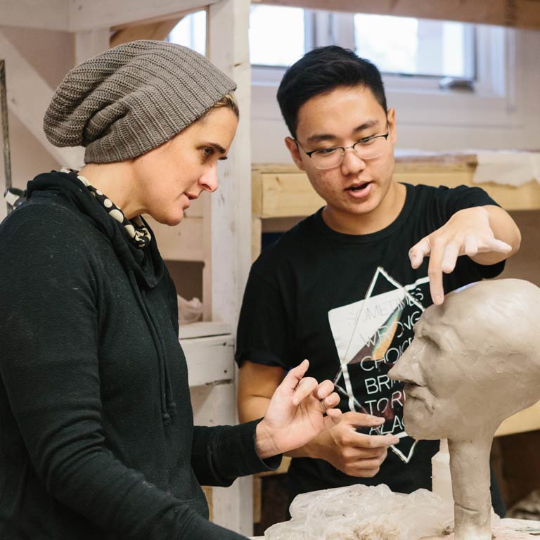 A professor works with a student on a sculpture of a face.