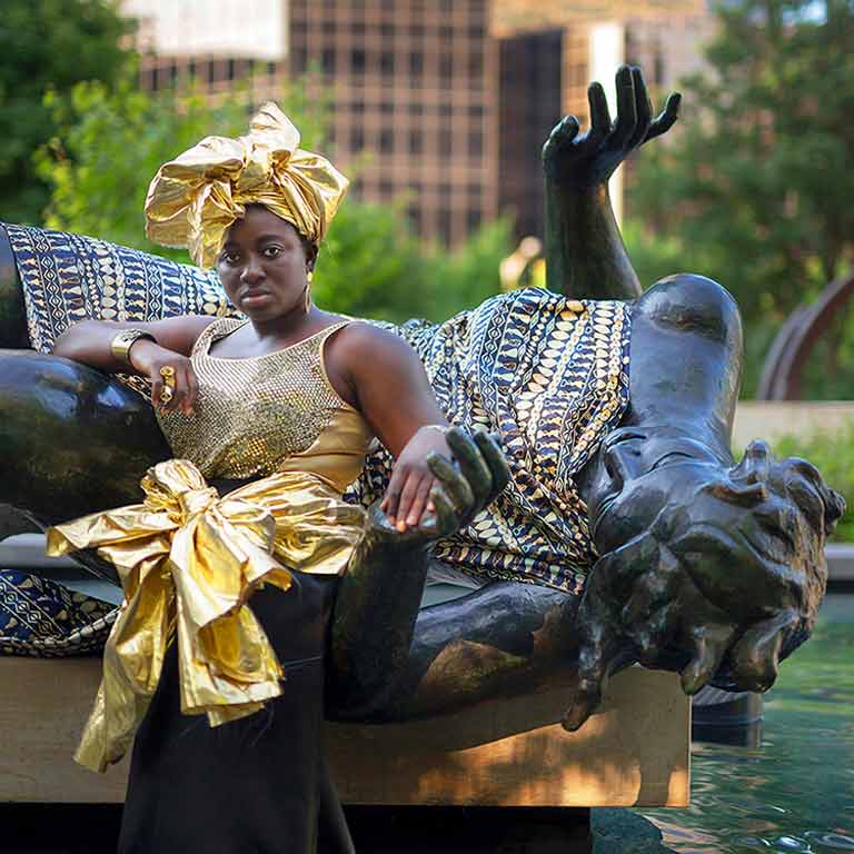 A person dressed in gold sits on a statue of a person laying down.