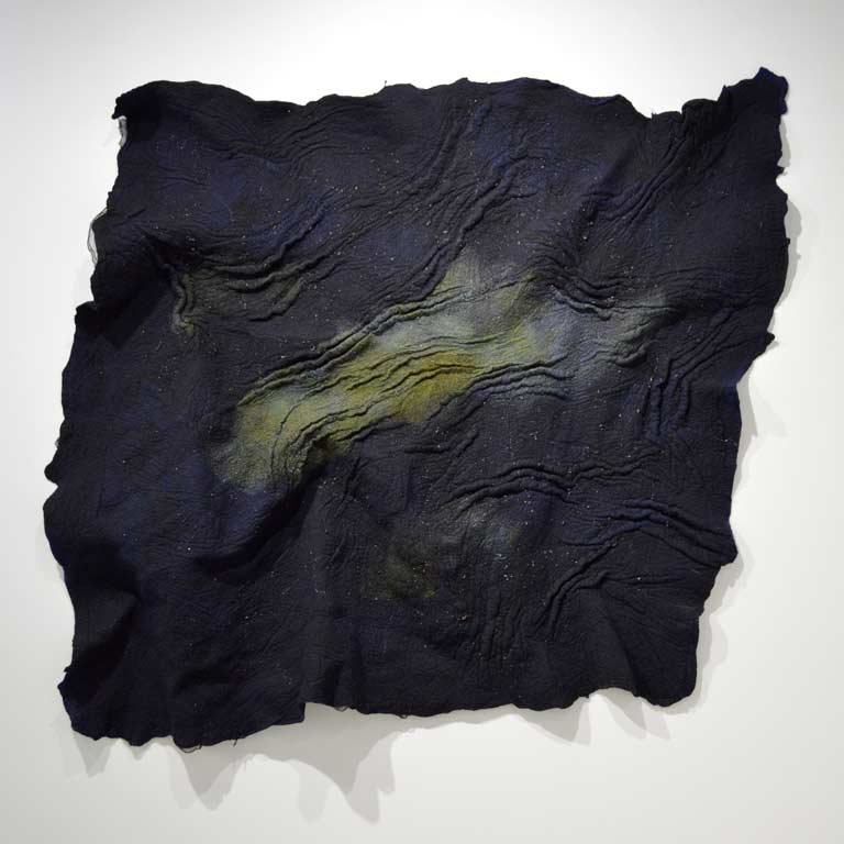 A navy blue fiber artwork hanging on a wall. It has various folds and waves set into it, culminating into a green center.