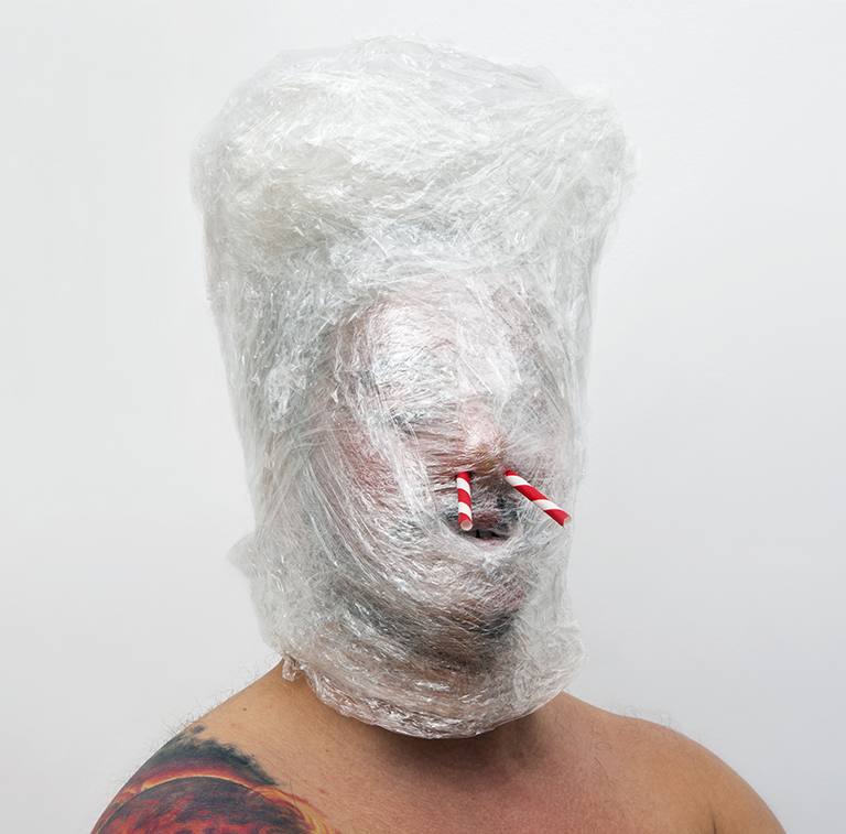 A sculpture of a face wrapped in saran wrap with straws in its nose. 