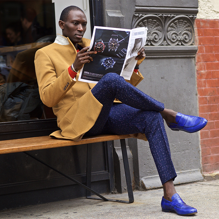 A well-dressed man sits on a bench reading the newspaper. 