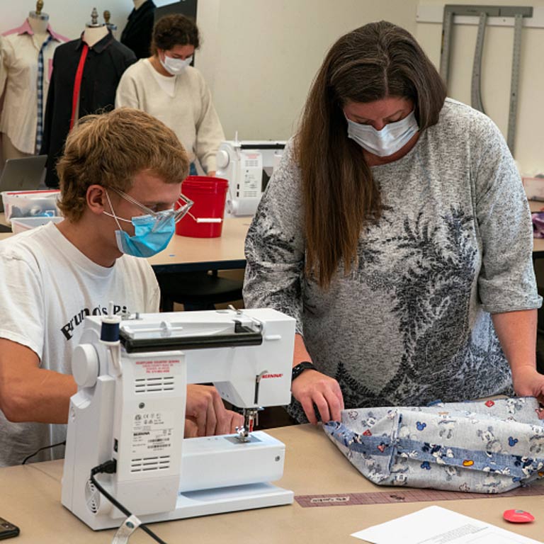 A professor takes a look at a students tote bag that they are sewing at a sewing machine.