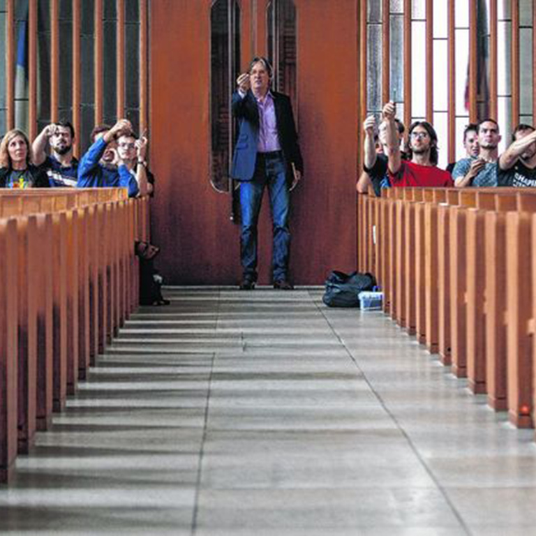 A person stands in a door holding something small in his hand. People seated in rows around him do the same thing.
