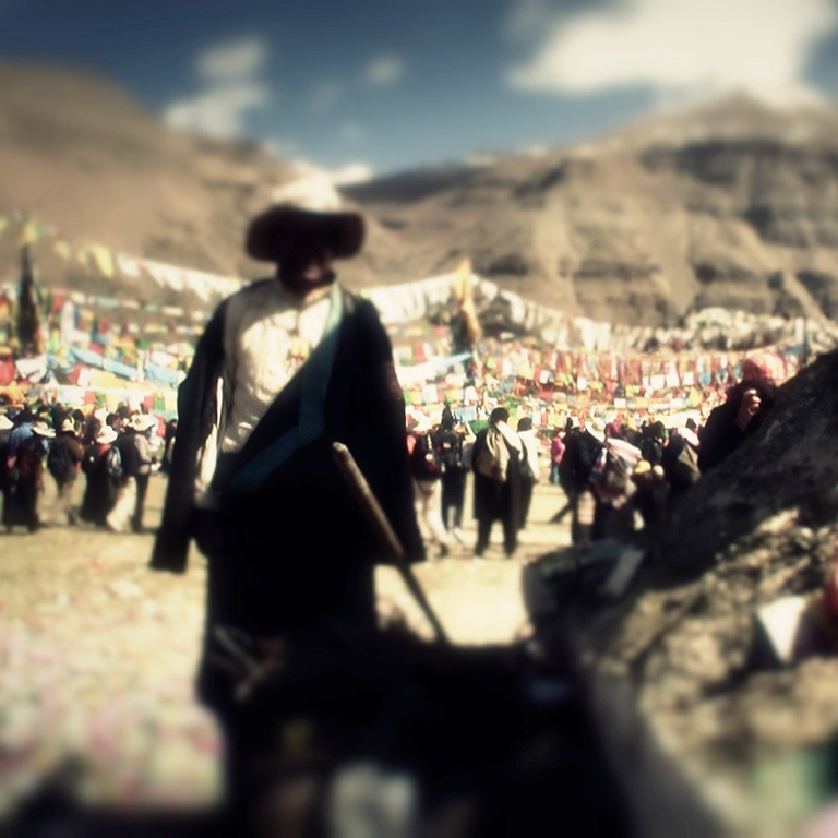 An out-of-focus photo of a man standing in front of a large group of people. A mountain is in the background