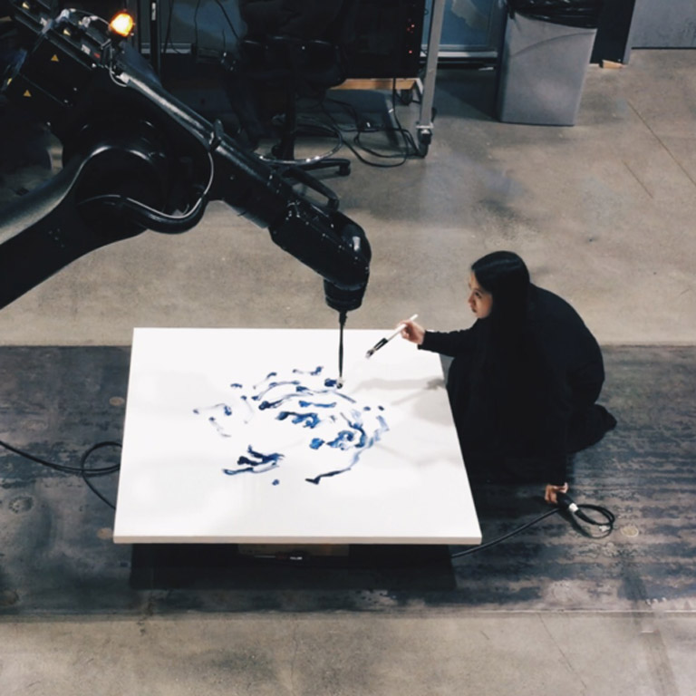 A person paints a canvas which is also being painted by a robot holding a paint brush